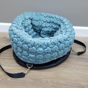May include: A light blue quilted pet bed with a black strap and a silver buckle. The bed is designed to be portable and can be carried by the strap.