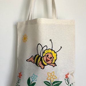 May include: A white canvas tote bag with a cartoon bee design. The bee is yellow and black with a smiling face and is flying over green grass and flowers. The bag has a green polka dot pattern at the bottom.