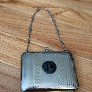 May include: A rectangular silver-tone metal cigarette case with a hinged lid and a decorative chain handle. The front features vertical etched stripes and a central circular black inlay depicting a silhouette of two figures.
