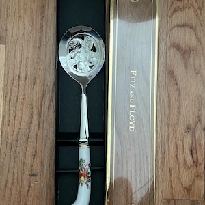 May include: A silver-toned serving spoon with a decorative floral design on the spoon head. The handle is white with a painted Christmas-themed design. The spoon is in a black box with a clear lid, and the box is labeled "FITZ AND FLOYD".