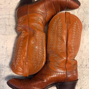 Puede incluir: Un par de botas vaqueras de cuero color canela con un patrón texturizado de piel de lagarto en el pie, una caña de cuero liso con intrincadas costuras decorativas claras, tiradores en la parte superior y un tacón de bloque marrón oscuro.