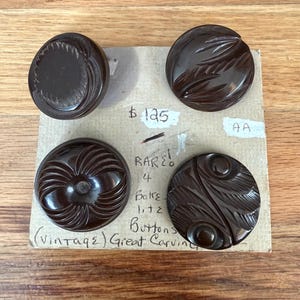 May include: Four dark brown circular buttons with intricate carved patterns attached to a rectangular cardboard backing. The card features handwritten text including $125, AA, RARE!, 4 Bakelite Buttons, and (vintage) Great Carving.