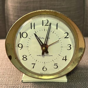 Peut inclure: Un réveil vintage doré Westclox Baby Ben avec un cadran blanc et des chiffres noirs. L'horloge a une base crème et indique "MADE IN USA". Les aiguilles indiquent environ 10h58.