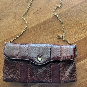 Pode incluir: Uma bolsa clutch retangular com padrão de pele de cobra em tons de castanho escuro e preto com acabamento brilhante. A bolsa possui uma aba dobrável fixada por um fecho central de metal dourado e uma alça de corrente metálica dourada.