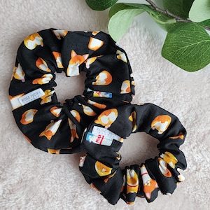 May include: Two black scrunchies with a white and orange print of cartoon corgis. The scrunchies have a white label that says "Rose Ivory Studio".