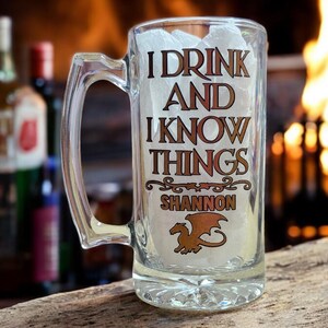 May include: A clear glass beer mug with a handle, featuring the text "I drink and I know things" in gold lettering. The mug also has a gold dragon design and the name "Shannon" printed on it.