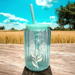 May include: A clear glass tumbler with a light blue lid and a white floral design. A light blue straw is inserted through the lid.