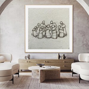 May include: A framed black and white line drawing of a group of people huddled together. The drawing is on a white background and is framed in a simple white frame. The drawing is hanging on a wall in a living room setting.