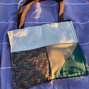 May include: A patchwork tote bag with brown handles. The bag features a variety of fabrics, including a green paisley print, tan, and a light beige gingham pattern. The bag is sitting on a purple striped surface.