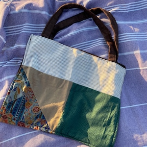 May include: A patchwork tote bag with brown handles, resting on a purple and white striped towel. The bag features panels of beige gingham, tan, green, and patterned fabric with floral and paisley designs. The bag is designed for carrying items.