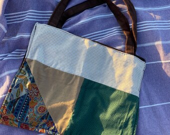 May include: A patchwork tote bag with brown handles, resting on a purple and white striped towel. The bag features panels of beige gingham, tan, green, and patterned fabric with floral and paisley designs. The bag is designed for carrying items.