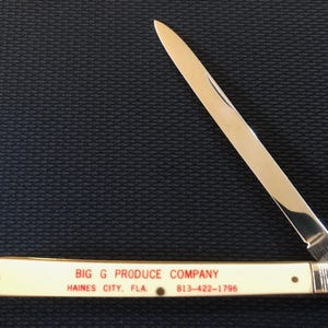 May include: A vintage pocket knife with a cream-colored handle and a silver blade. The handle features red text that reads "BIG G PRODUCE COMPANY, HAINES CITY, FLA. 813-422-1796". The blade is partially open, revealing its sharp edge.