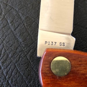 May include: Close-up of a partially visible knife. The stainless steel blade has the text "P137 SS" etched on it. The handle is made of wood and has a brass-colored rivet. The background is a textured black surface.