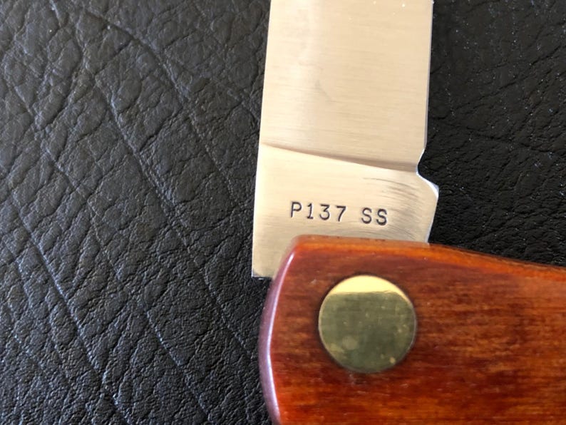 May include: Close-up of a partially visible knife. The stainless steel blade has the text "P137 SS" etched on it. The handle is made of wood and has a brass-colored rivet. The background is a textured black surface.