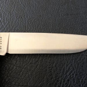 May include: Close-up of a stainless steel knife blade with a wooden handle. The blade is shiny and has the text "P137 SS" etched near the handle. The knife is set against a dark, textured background.