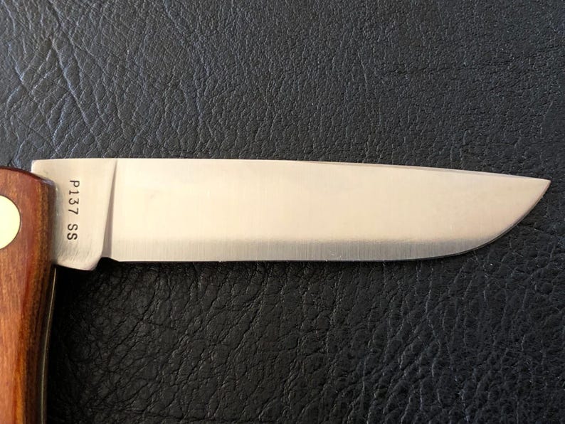 May include: Close-up of a stainless steel knife blade with a wooden handle. The blade is shiny and has the text "P137 SS" etched near the handle. The knife is set against a dark, textured background.