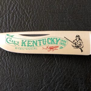 May include: A stainless steel Case XX pocket knife blade with a wood handle. The blade features the text "Case's Kentucky Bicentennial" in green, with a red bird graphic and a drawing of a person holding a rifle.