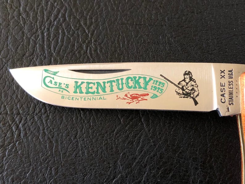 May include: A stainless steel Case XX pocket knife blade with a wood handle. The blade features the text "Case's Kentucky Bicentennial" in green, with a red bird graphic and a drawing of a person holding a rifle.