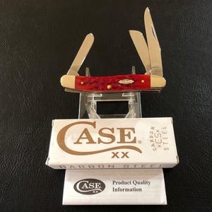 May include: A red and gold Case XX pocket knife with three open blades. The knife rests on a clear stand above a white box and a product information sheet. The box has the Case XX logo and the words "CARBON STEEL".