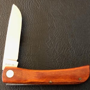 May include: A folding pocket knife with a wooden handle and a partially open silver blade. The handle is a rich brown color, and the blade is marked with "P137 SS". The knife is set against a dark, textured background.