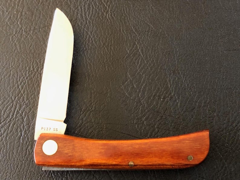May include: A folding pocket knife with a wooden handle and a partially open silver blade. The handle is a rich brown color, and the blade is marked with "P137 SS". The knife is set against a dark, textured background.