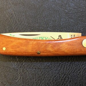 May include: A wooden-handled pocket knife with a partially visible blade. The handle is a warm brown color with brass rivets. The blade has text and an illustration. The knife is closed and resting on a dark surface.