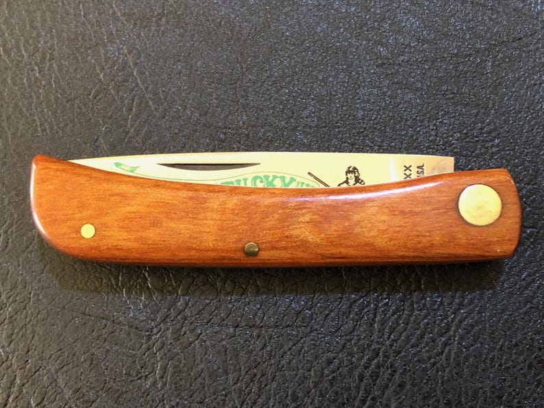 May include: A wooden-handled pocket knife with a partially visible blade. The handle is a warm brown color with brass rivets. The blade has text and an illustration. The knife is closed and resting on a dark surface.