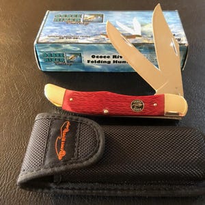 May include: A red folding hunting knife with a gold bolster and three blades, next to a black sheath and a box. The knife has the text "Ocoee River" on the handle and the box.