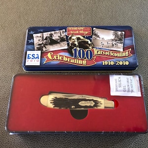 May include: A Schrade Uncle Henry pocket knife presented in a red-lined tin. The tin lid features a blue design with the text "Celebrating 100 Years of Scouting 1910-2010" and historical images. The knife is in a protective case.