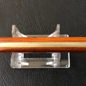 May include: A wooden knife with a brass accent, displayed on a clear acrylic stand. The knife has a dark brown handle and a light brown blade. The brass accent runs along the length of the knife.