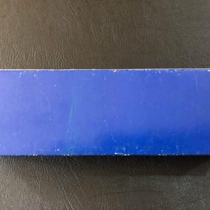 May include: A rectangular, blue box with white edges. The box is a solid, vibrant blue color. The surface of the box has some minor scratches and wear. The box is sitting on a dark, textured surface.