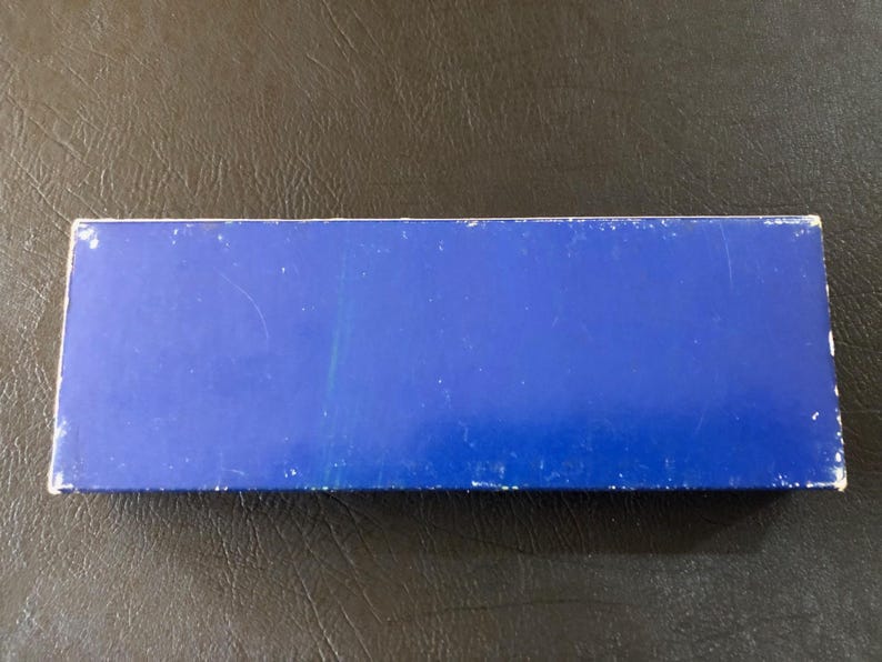 May include: A rectangular, blue box with white edges. The box is a solid, vibrant blue color. The surface of the box has some minor scratches and wear. The box is sitting on a dark, textured surface.