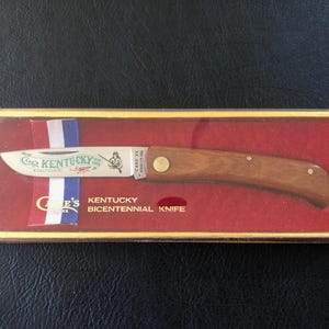 May include: A Case XX Kentucky Bicentennial Knife in a gold-trimmed box. The knife has a wooden handle and a silver blade with the words "Kentucky" and an illustration. The box has red and white, blue ribbon.