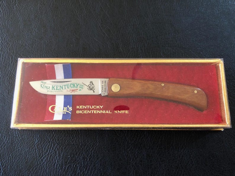 May include: A Case XX Kentucky Bicentennial Knife in a gold-trimmed box. The knife has a wooden handle and a silver blade with the words "Kentucky" and an illustration. The box has red and white, blue ribbon.