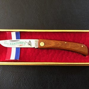 May include: A wooden-handled pocket knife with a stainless steel blade, presented in a gold-trimmed box with a red velvet interior. The blade features the word "Kentucky" and an illustration. A ribbon with red, white, and blue stripes is also in the box.