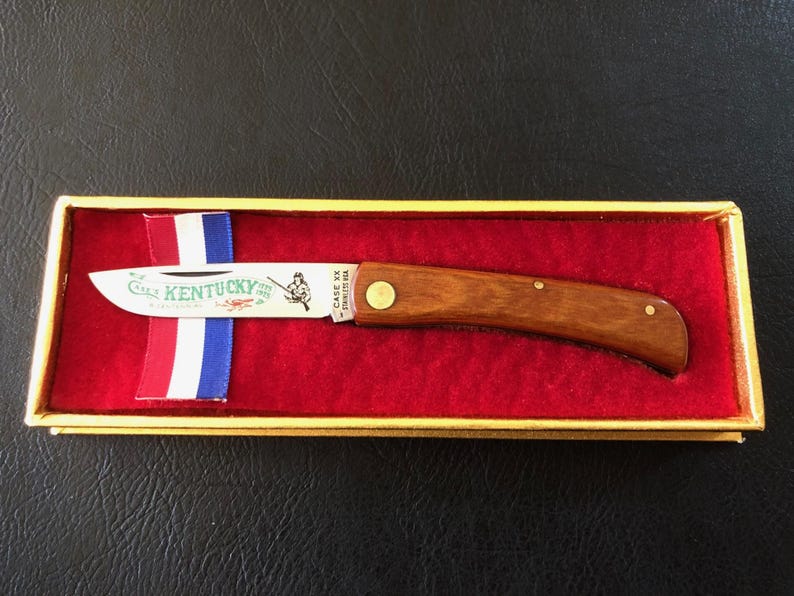 May include: A wooden-handled pocket knife with a stainless steel blade, presented in a gold-trimmed box with a red velvet interior. The blade features the word "Kentucky" and an illustration. A ribbon with red, white, and blue stripes is also in the box.