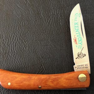 May include: A vintage folding knife with a wooden handle and a stainless steel blade. The blade features the text "Kentucky" and an illustration. The handle is a warm brown color, and the knife is closed in the image.
