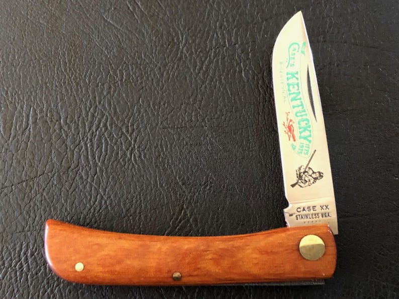May include: A vintage folding knife with a wooden handle and a stainless steel blade. The blade features the text "Kentucky" and an illustration. The handle is a warm brown color, and the knife is closed in the image.