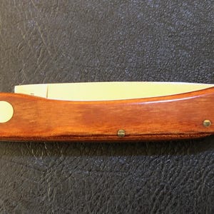 May include: A wooden folding knife with a light-colored blade. The handle is a rich, warm brown with a circular inlay and two brass rivets. The knife is closed, showing the full length of the handle and blade.