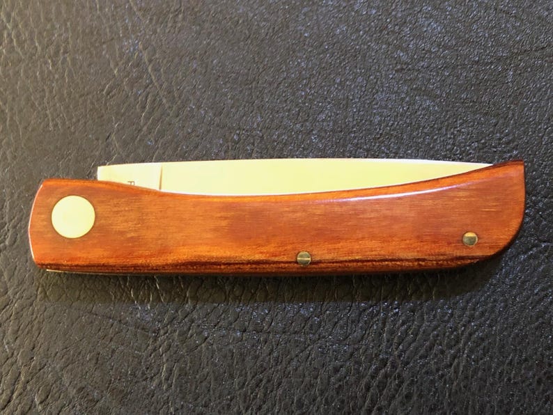 May include: A wooden folding knife with a light-colored blade. The handle is a rich, warm brown with a circular inlay and two brass rivets. The knife is closed, showing the full length of the handle and blade.