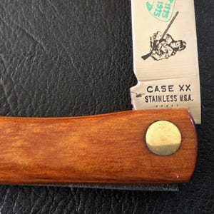 May include: Close-up of a Case XX stainless steel pocket knife. The knife has a wooden handle with a brass rivet. The blade is partially open, displaying an illustration and the text "CASE XX STAINLESS U.S.A."