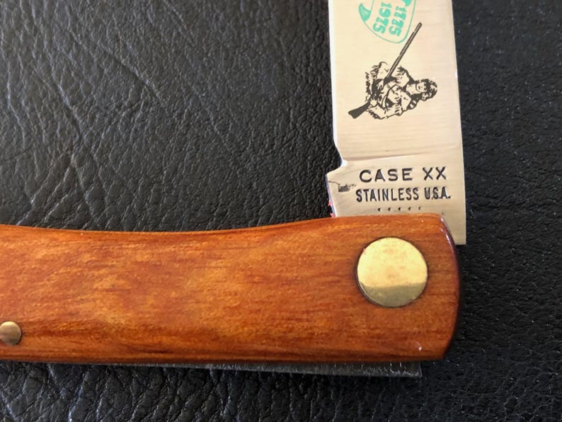 May include: Close-up of a Case XX stainless steel pocket knife. The knife has a wooden handle with a brass rivet. The blade is partially open, displaying an illustration and the text "CASE XX STAINLESS U.S.A."