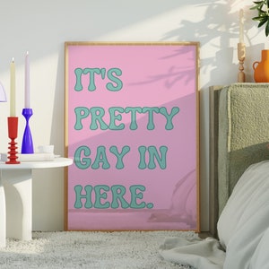 May include: A pink framed poster with the text "It's Pretty Gay In Here." The poster is hanging on a white wall above a bed with a green headboard. There are two candlesticks and a lamp on a white side table.