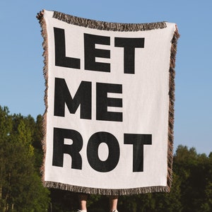 Peut inclure: Une personne tient une tapisserie tissée blanche avec du texte noir qui dit "LET ME ROT".