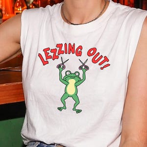 Può includere: T-shirt bianca senza maniche con la scritta "LEZZING OUT!" in rosso, sopra una grafica di una rana verde che tiene le forbici. La rana ha la pancia gialla. La persona indossa jeans blu e un braccialetto d'argento.