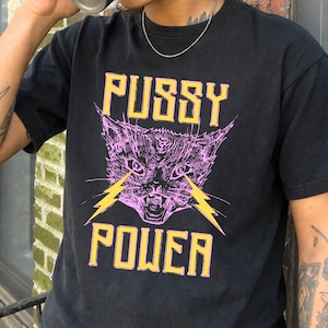 May include: Black crew neck t-shirt featuring a pink cat graphic with yellow lightning bolts and the words "Pussy Power". The design is printed on the front of the shirt.