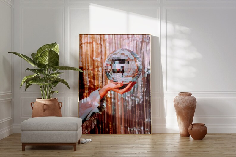 May include: A framed print of a hand holding a silver disco ball against a shimmering background of gold and pink.