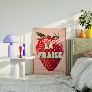 K&ouml;nnte beinhalten: Ein rosa-wei&szlig;es Poster mit einer gro&szlig;en roten Erdbeerillustration und dem Text "LA FRAISE". Das Poster h&auml;ngt an einer wei&szlig;en Wand &uuml;ber einem Bett mit einem gr&uuml;nen Kopfteil.