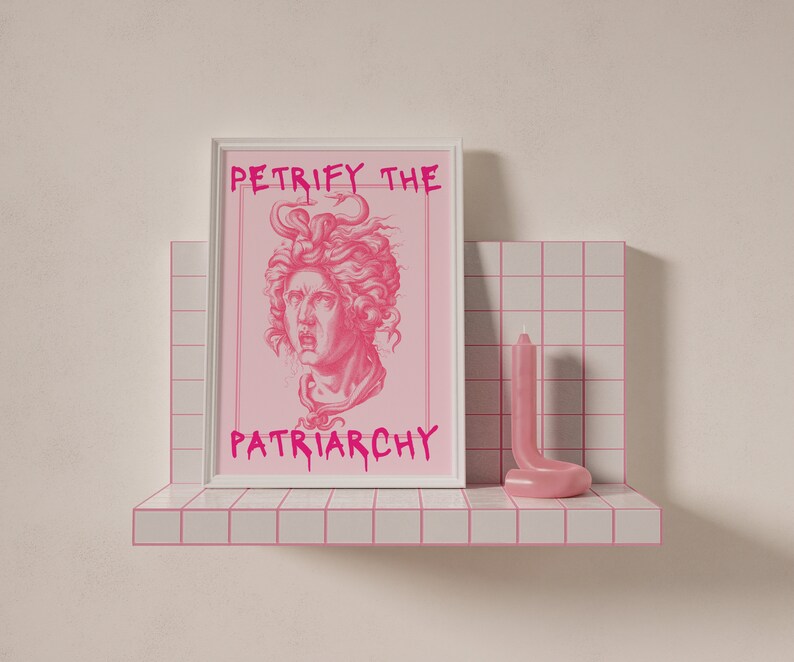May include: A white framed print with the text "Petrify the Patriarchy" in pink lettering. The print features a pink illustration of a woman's face with curly hair. The print is on a shelf with a pink candle.