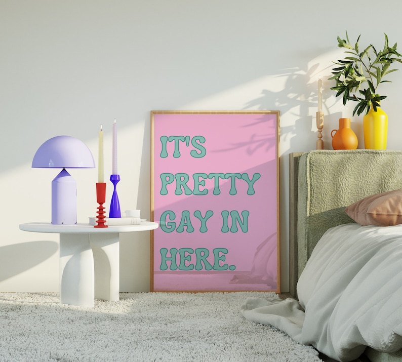 May include: A pink framed poster with the text "It's Pretty Gay In Here." The poster is hanging on a white wall above a bed with a green headboard. There are two candlesticks and a lamp on a white side table.